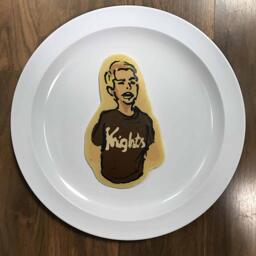 An image of a pancake drawn in the shape of a young man wearing a 'Knights' T-shirt with his mouth open in a funny expression.