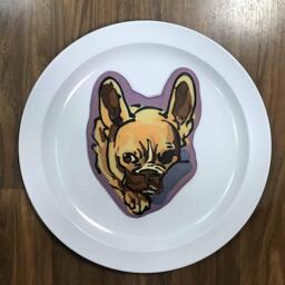 An image of a pancake drawn in the shape of a cute french bulldog, hiding its nose behind a blanket.