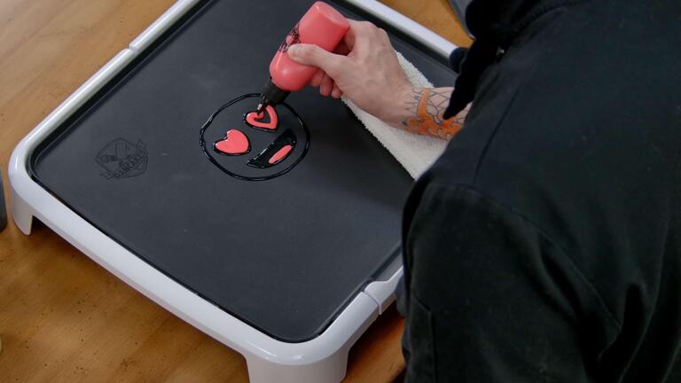 An image showing the black outlines of a heart eyes emoji pancake design being filled in with red batter, in the mouth adn heart-shaped eyes.