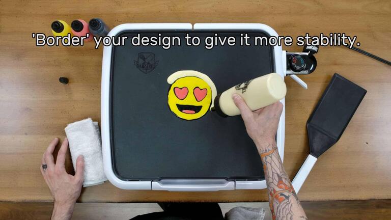 An image of a completed heart-eyes emoji pancake design, face down on the griddle, while the artist uses their fill bottle to add a thick border of plain batter around the outside of this design. The image reads, "'Border' your design to give it more stability."