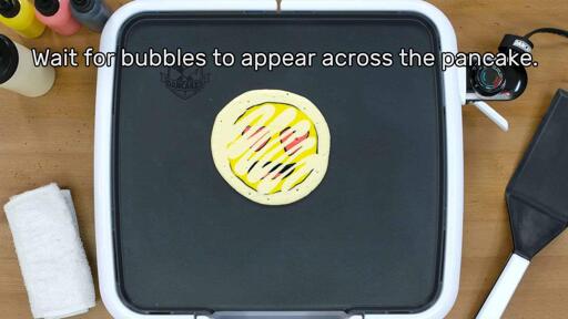 An image of a heart-eyes emoji pancake design that has been bordered and backed, beginning to bubble on the griddle as it cooks. The image reads