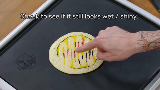 This image shows the artist pointing to the back of the cooking pancake art design to point out how the texture of the pancake is slowly changing from shiny to matte. As it cooks, it becomes less 'wet'. The image reads
