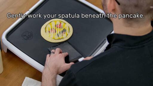 This image shows the artist, holding the spatula, starting to slide the spatula under and around the pancake design, which is gently stuck in place. The image reads