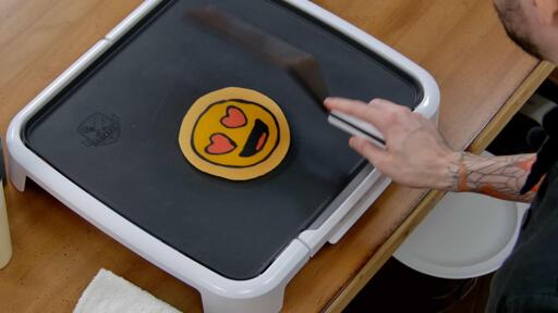 An image that shows the heart eyes emoji pancake design hit the griddle as it is flipped over, revealing its final appearance. The spatula is in frame, with a motion blur.