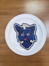 Pancake art of a midnight blue anthro/furry character with their tongue sticking out, blue sideburns, and big, shiny purple eyes and white horns on the top of their head. A stark outline of white batter makes the design pop.