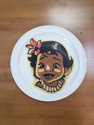 Pancake art of baby Moana from the animated film, Moana. She looks curious and excited, and has a bright pink flower in her hair.