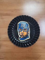 Pancake art of Captain America, wearing his blue hood/cap with a large white 'A' emblazoned across. He has a disgruntled facial expression.