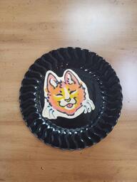 Pancake art of a simple cartoon cat with orange fur, a white snout, and yellow highlights. The cat is smiling with its tongue out and its eyes closed, and looks content.