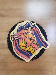 Pancake art of a red dragon with a vicious scowl and white eyes with no pupils. Its chest scales are golden yellow and the skin of its wings are a deep purple, and it has two curved horns atop its head.