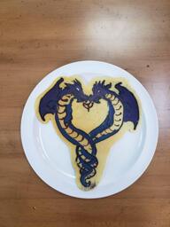 Pancake art of two purple serpent dragons with single winges entwined in the shape of a heart. A small spit of flame comes from each of their mouths to create the point in the center of the heart.