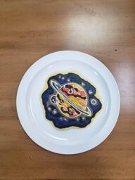 Pancake art of a simple abstact space scene, in which a large planet with an atmosphere of roiling reds, oranges, browns and yellows (with simple rings around it like Saturn) sits against a black backdrop with a few stars, two smaller moons, and a blue/purple mixture of nebulous space gasses giving is some mysterious character.