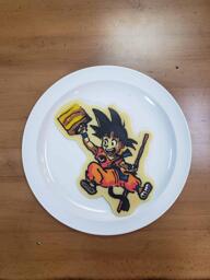 Pancake art of a young Son Goku (from the dragonball anime) holding his staff and carrying a suitcase, smiling as though he is about to go on vacation. He has his monkey tail extended beneath him.