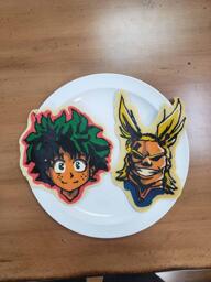 Pancake art of Midoriya and All Might from the popular anime My Hero Academia. Midoriya has an unkempt tousle of dark green hair and a bright, open expression of contenment, and All Might has a dramatic hairstyle with two arcing yellow locks almost like rabbits ears or antennae, and his prideful, mighty facial expression with his wide, full-teethed smile, is cast in heavy shadow, as though he is looking down on the viewer from a high, well-lit place.