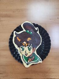 Pancake art of Sailor Saturn, from the Sailor Moon anime. Sailor Saturn is a young woman with a golden tiara-type headdress resting on her forehead underneath a thick coif of long, dark brown hair, with two green beads tying her hair into a ponytail behind her. She has a green chocker and a green bandana with two white stripes towards it, and is smiling with large, shining green irises.