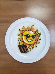 Pancake art of a yellow sun with sunglasses, casually saluting and carrying a brown suitcase as if it is about to go on vacation. It has a cheeky smile.