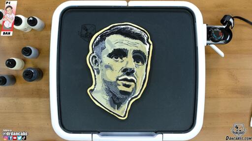 An image of a pancake art portrait of social media personality Gary Vaynerchuck, rendered in grayscale and pictured resting on the dancakes griddle from a top-down viewing angle. The warm brown wood of the countertop and the multicolored batter pens adorn the edges of this simple image.